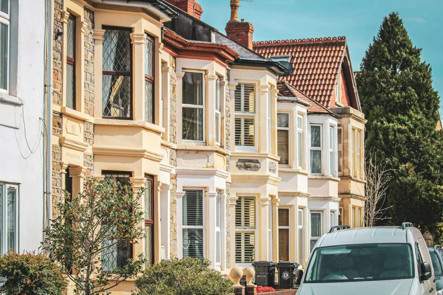 Row of Victorian terraced houses with cream painted bay windows and stone facades, small front gardens with shrubs, and a white van parked in the foreground on a sunny day; no readable text; calm and welcoming residential scene.